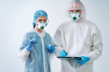 Doctor holds clipboard and nurse holds an injection syringe and vaccine. Healthcare workers in protective suits and medical masks