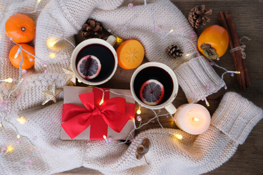 two cups of hot mulled wine with lemon, persimmon and tangerine fruits, candles, box with gift in kraft paper, with red bow lies on table, concept of holiday, congratulations merry christmas, new year