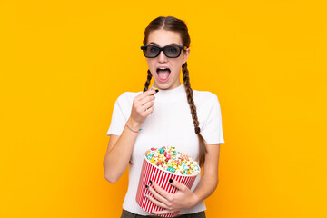 Obraz premium Young woman over isolated yellow background with 3d glasses and holding a big bucket of popcorns