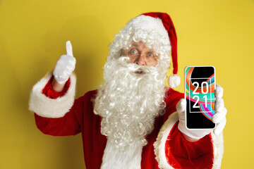 Stylish Santa Claus in traditional costume with modern device pointing, showing on 2021 on the screen on yellow studio background. Technologies, celebration, winter mood and emotions concept