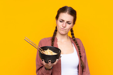 Young woman over isolated yellow background with sad expression while holding a bowl of noodles...