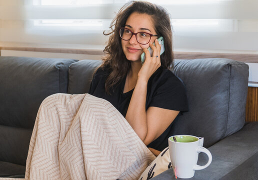 Hispanic Woman Sitting On A Sofa With A Blanket And Cup Of Tea While Talking To The Phone