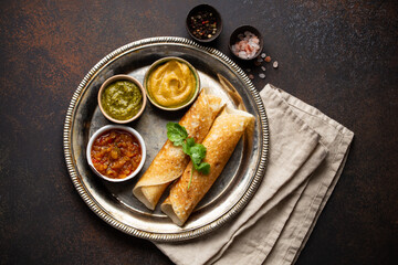 Traditional Indian rice pancakes Dosa with different dips chutney, seasonings on rustic metal plate on dark brown stone background table. Quick meal or vegetarian snack of South India, top view