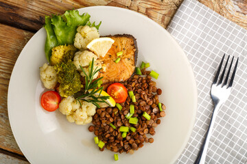 Baked fish with broccoli and lentils in a white plate with rosemary and lemon on a napkin next to a fork.