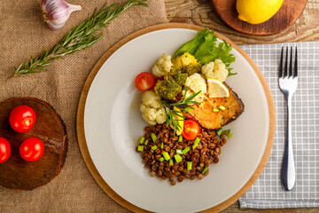 Baked fish with broccoli and lentils in a white plate with rosemary and lemon on a round stand next to a fork and vegetables.