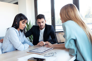Fototapeta premium Beautiful real-estate agent showing house plans on electronic tablet while talking to the couple about buying the house in the office.