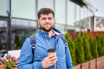 Bearded man with cup of takeaway coffee stands outdoors.