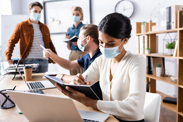 african american businesswoman in medical mask writing in notebook near colleagues working on blurred background