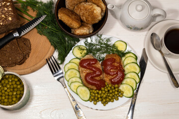 On a white plate are cutlets with tomato sauce, green peas, pieces of cucumber and a sprig of dill. Nearby is a tea set, a board of bread, a bowl of cutlets, and a jar of peas.