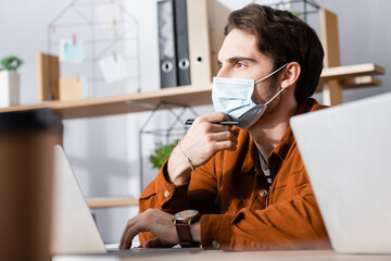thoughtful businessman in medical mask looking while holding pen on blurred foreground