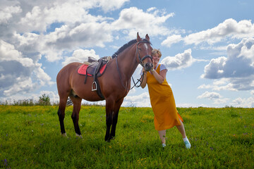 Girl in yellow dress with a horse on a green field and a blue sky with white clouds on the background
