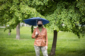 Woman in a black mask under an umbrella in the rain. Woman in a brown jacket and khaki pants. Green trees and grass in the background.