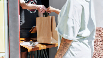 Cropped shot of woman taking paper bag with her order from hands of shop assistant while collecting...