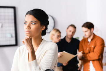 pensive african american businesswoman looking away while multiethnic managers talking on blurred background