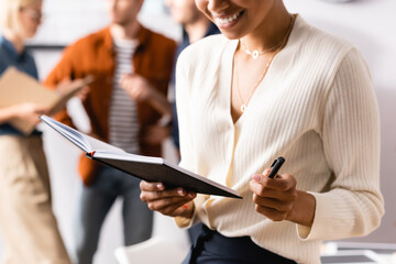 cropped view of african american businesswoman holding notebook and pen near colleagues on blurred background