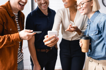 cheerful businesspeople pointing with finger and pen and smartphone near multicultural colleagues on blurred background