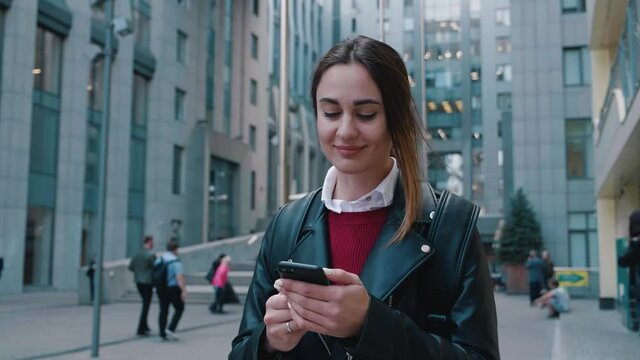 A Young Woman Stands With A Phone On The Background Of A Business Center. Online service taxi order mobile application. 4k
