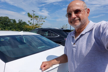 A smiling mature bald Caucasian man with his arm on the trunk of a white car