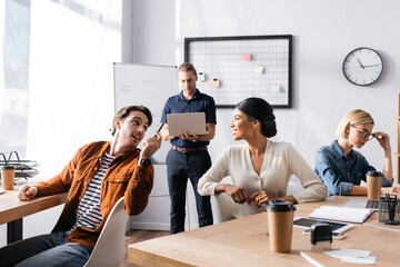 Smiling multicultural office workers talking while sitting at tables in office with colleagues on background