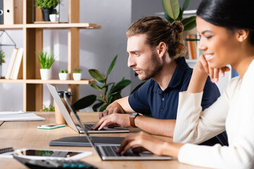 Focused young adult man looking at laptop while sitting at workplace with blurred african american woman on foreground