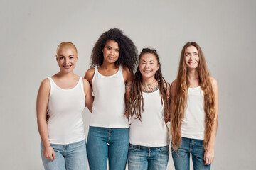 Portrait of four young diverse women wearing white shirts and denim jeans smiling at camera while posing together isolated over grey background