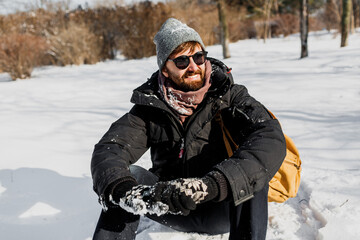 Winter Lifestyle Portrait of handsome man with beard and backpack  sitting on snow in winter...