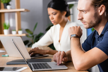 Focused office worker with watch using laptop while sitting at workplace with blurred african american woman on background