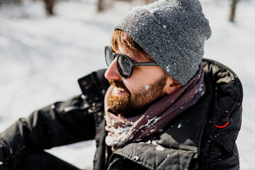 Close up winter portrait of hipster man with beard in grey hat relaxing in  sunny park with snowflakes on clothes.