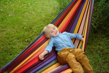 Fototapeta na wymiar Cute little blond caucasian boy having fun with multicolored hammock in backyard or outdoor playground. Summer active leisure for kids. Child swinging and relaxing in hammock.