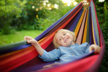 Fototapeta na wymiar Cute little blond caucasian boy enjoy and having fun with multicolored hammock in backyard or outdoor playground. Summer active leisure for kids. Child swinging and relaxing in hammock.
