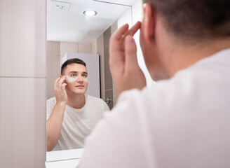 Man look in bathroom mirror use under eye anti-wrinkle patches, millennial male do morning facial procedures with hydrogel beauty product, skincare concept