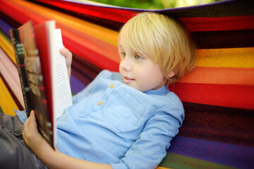 Fototapeta na wymiar Cute little blond caucasian boy reading book and having fun with multicolored hammock in backyard or outdoor playground. Summer leisure for kids. Child swinging and relaxing in hammock.