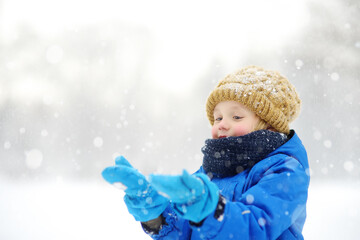 Little boy having fun playing with fresh snow during snowfall. Admiring baby catching snowflakes on...