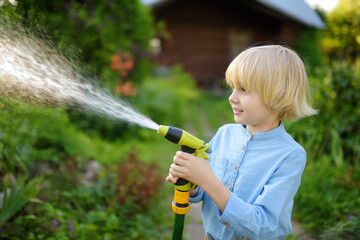 Funny little boy watering plants and playing with garden hose with sprinkler in sunny backyard....