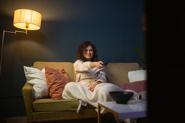 Young beautiful girl is sitting on the sofa and watching TV. The woman switches channels on the TV. A young woman is watching television series.