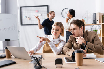 Focused man sitting at workplace near woman pointing with finger, while using laptop with multicultural office workers on background