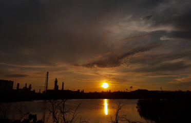 the sun sets over the lake and among the factories and trees