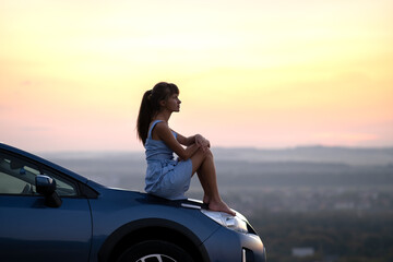 Happy young woman driver in blue dress leaning on her car hood enjoying warm summer day. Travelling and vacation concept.