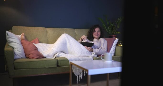 A modern young woman lies on the couch watching a movie and laughing. A girl with popcorn lies on the couch late at night watching TV shows. TV series, movies.