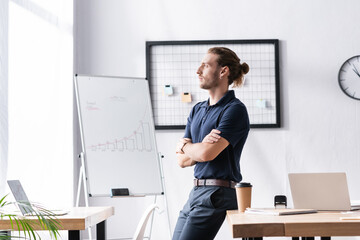 Confident businessman with crossed arms looking away while leaning on table in office