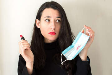 Una mujer joven morena sujeta una mascarilla quirúrgica azul manchada de pintalabios con una mano...