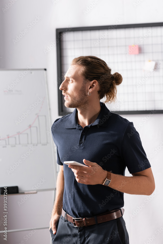 Wall mural Confident businessman with smartphone looking away while standing in office on blurred background