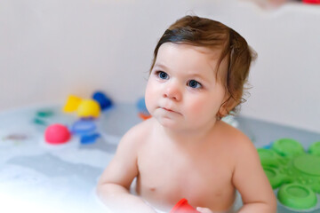 Cute adorable baby girl taking foamy bath in bathtub. Toddler playing with bath rubber toys.