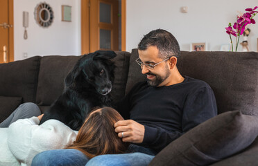 Young happy couple on the sofa at home with their black dog. Woman resting her head on the legs of her partner.