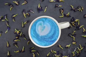 Blue tea latte in cup with heart shape, dried butterfly pea flowers pattern on gray concrete background. Menu, recipe, beverage concept. Flat lay, top view