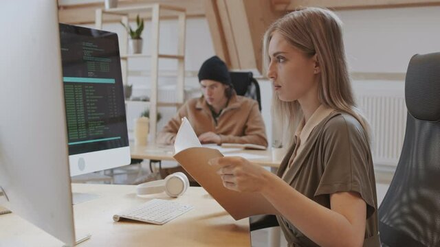 Pan shot of young blond-haired Caucasian woman wearing casual clothes sitting at desk and comparing code on computer screen with data in notebook in her hands