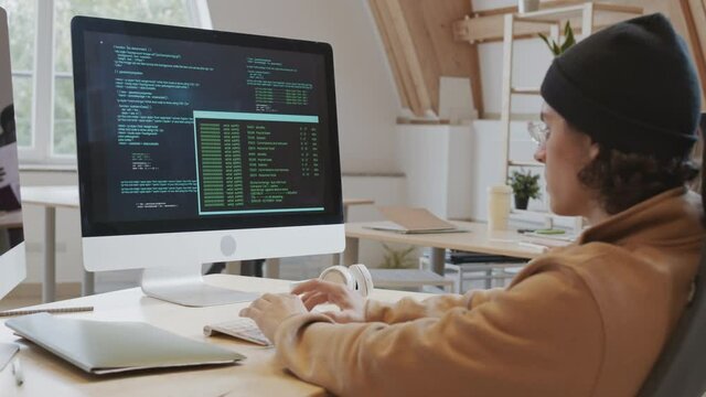 Back view of young Caucasian male coder wearing hoody, hat and glasses sitting at workplace and developing program code on computer