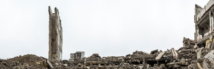 Panorama. The remains of a building with piles of gray concrete rubble and a detached ruined wall against a hazy sky.
