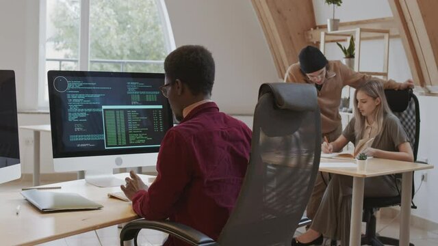 Back view of young African male coder wearing casual clothes sitting at workplace and developing program code on computer while his colleagues talking
