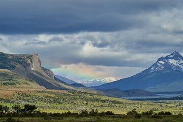 vast open landscape in Patagonia with dramatic sky and a rainbow over a valley, snow covered mountains of the Andes in the background